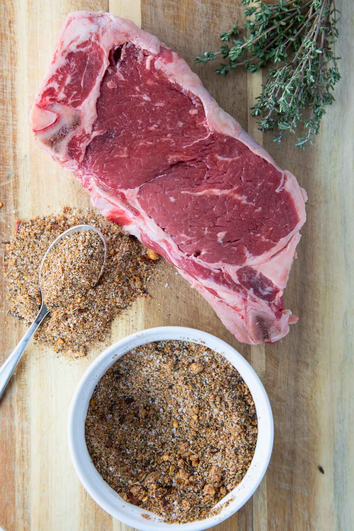 overhead of bowl of easy steak seasoning, steak seasoning with spoon, steak, and greens on a wooden cutting board